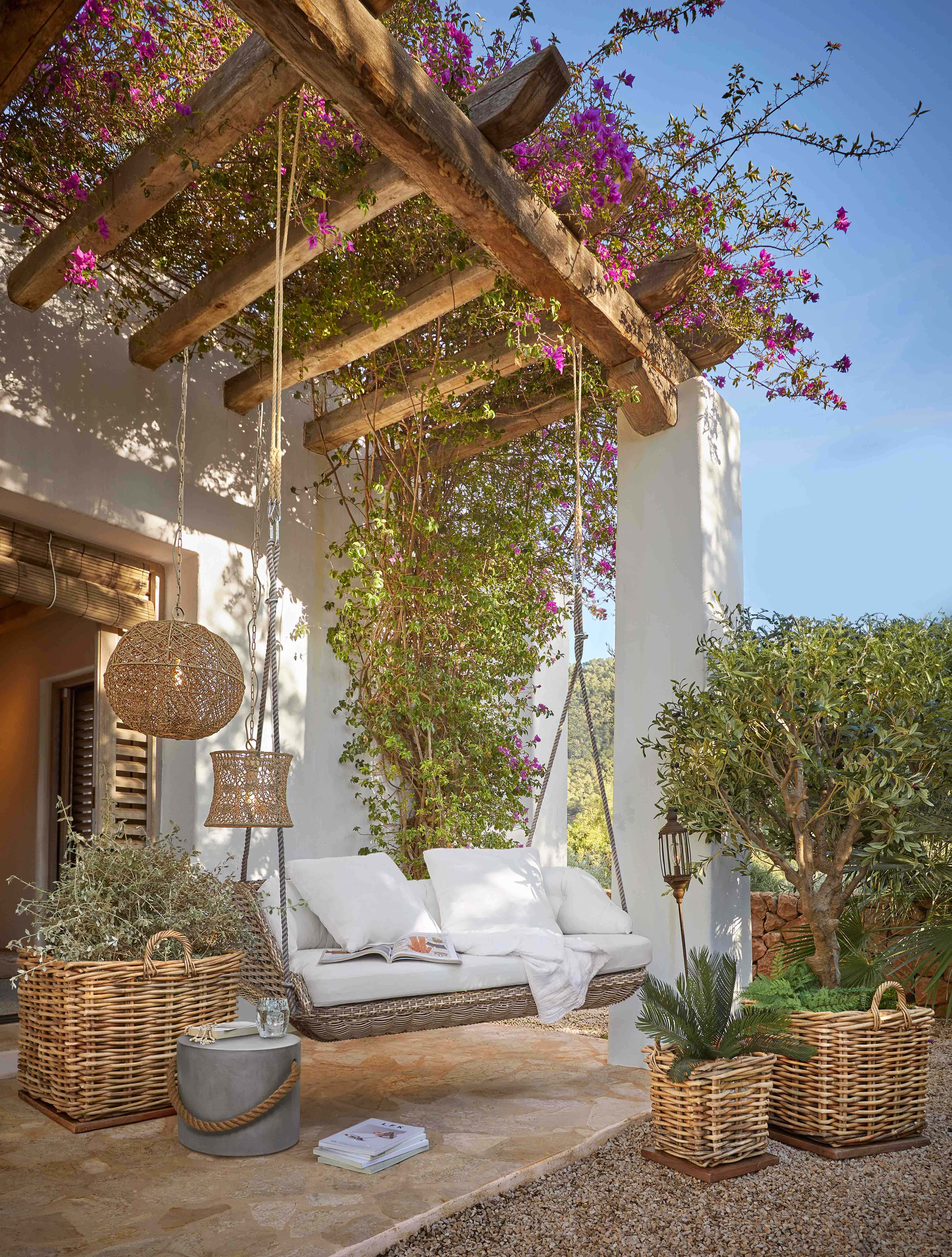 Huśtawka z rattanu z białymi poduszkami pod drewnianą pergolą z kwitnącymi różowymi kwiatami