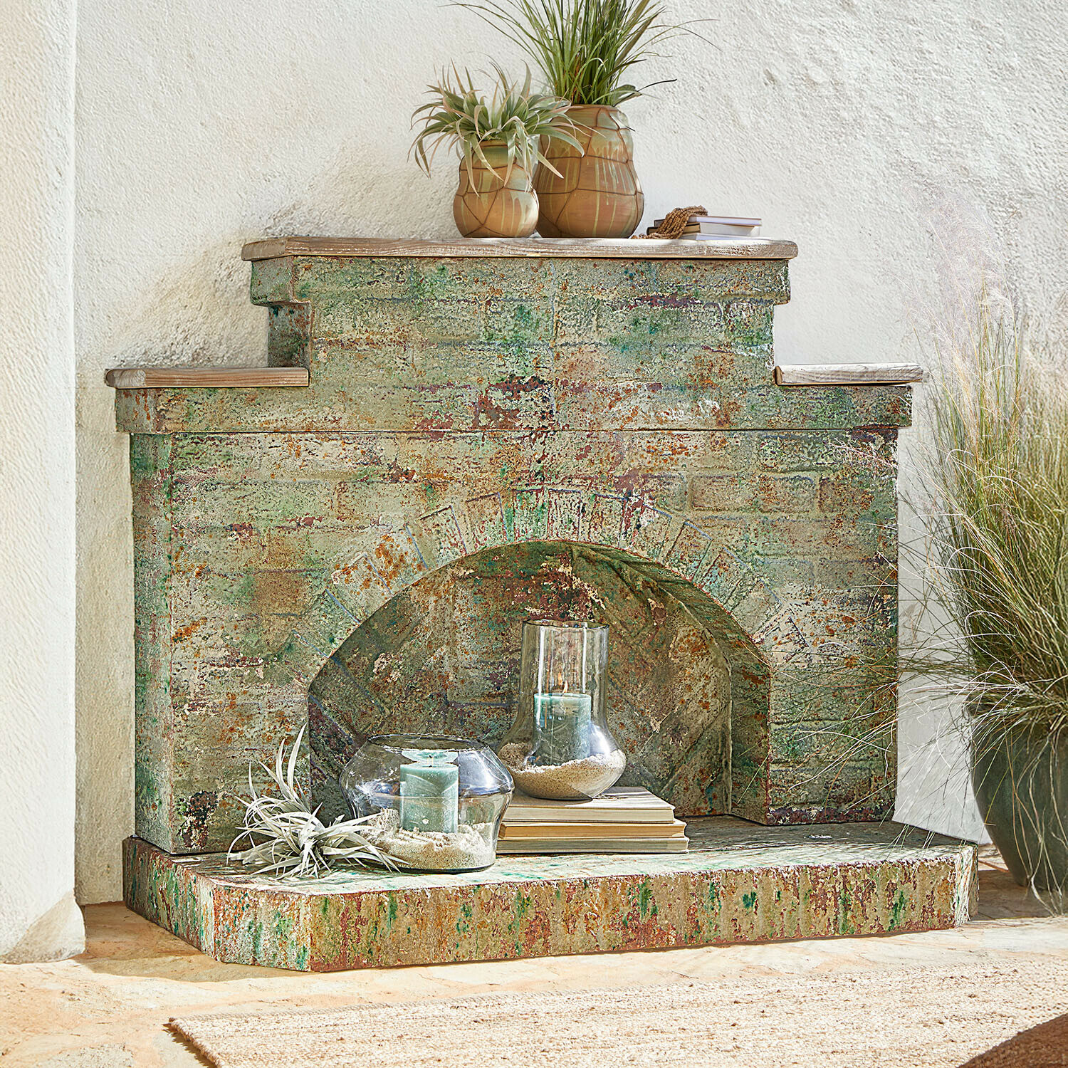 Dekoracyjny kominek z postarzanego metalu w zielono-rdzawych odcieniach z świecami i roślinami
