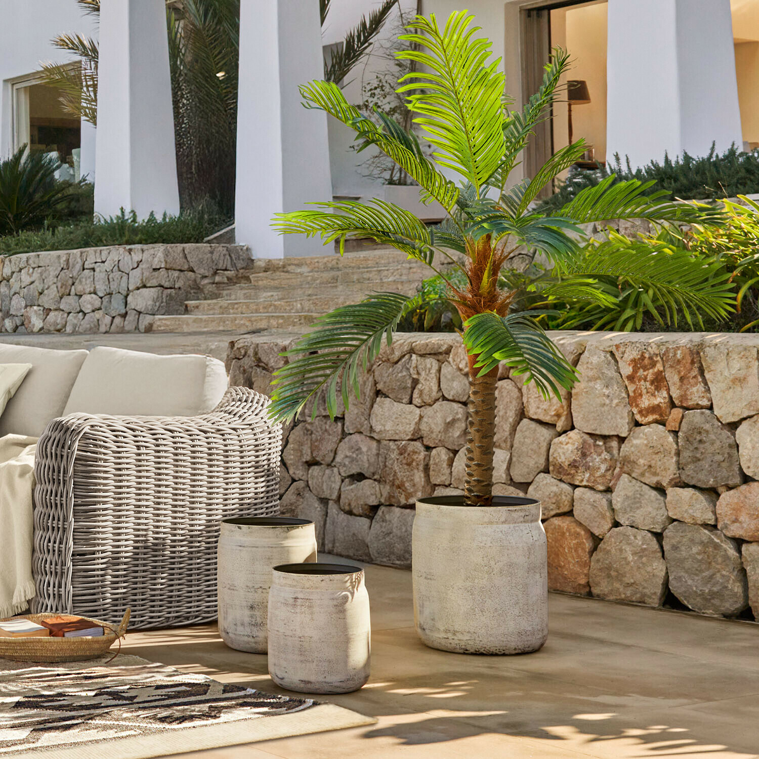 Zestaw trzech donic z jasnego betonu na taras z zieloną sztuczną palmą i meblami z rattanu