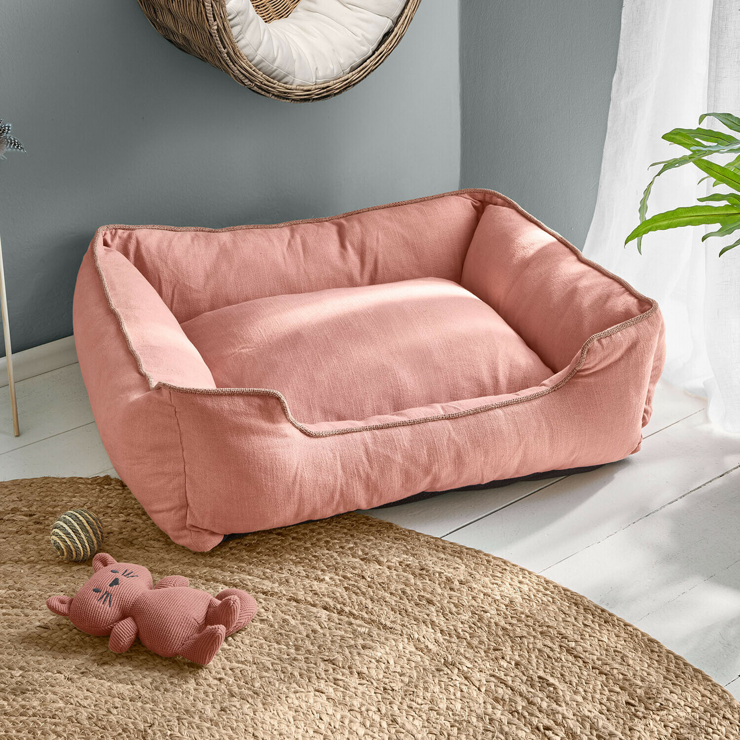 Różowe legowisko dla psa z miękkiego materiału na jasnej podłodze z zabawką kota i dywanem z juty