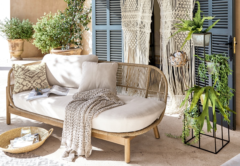 Boho kanapa z jasnym beżowym obiciem i drewnianą ramą, dekorowana poduszkami i plecionym kocem