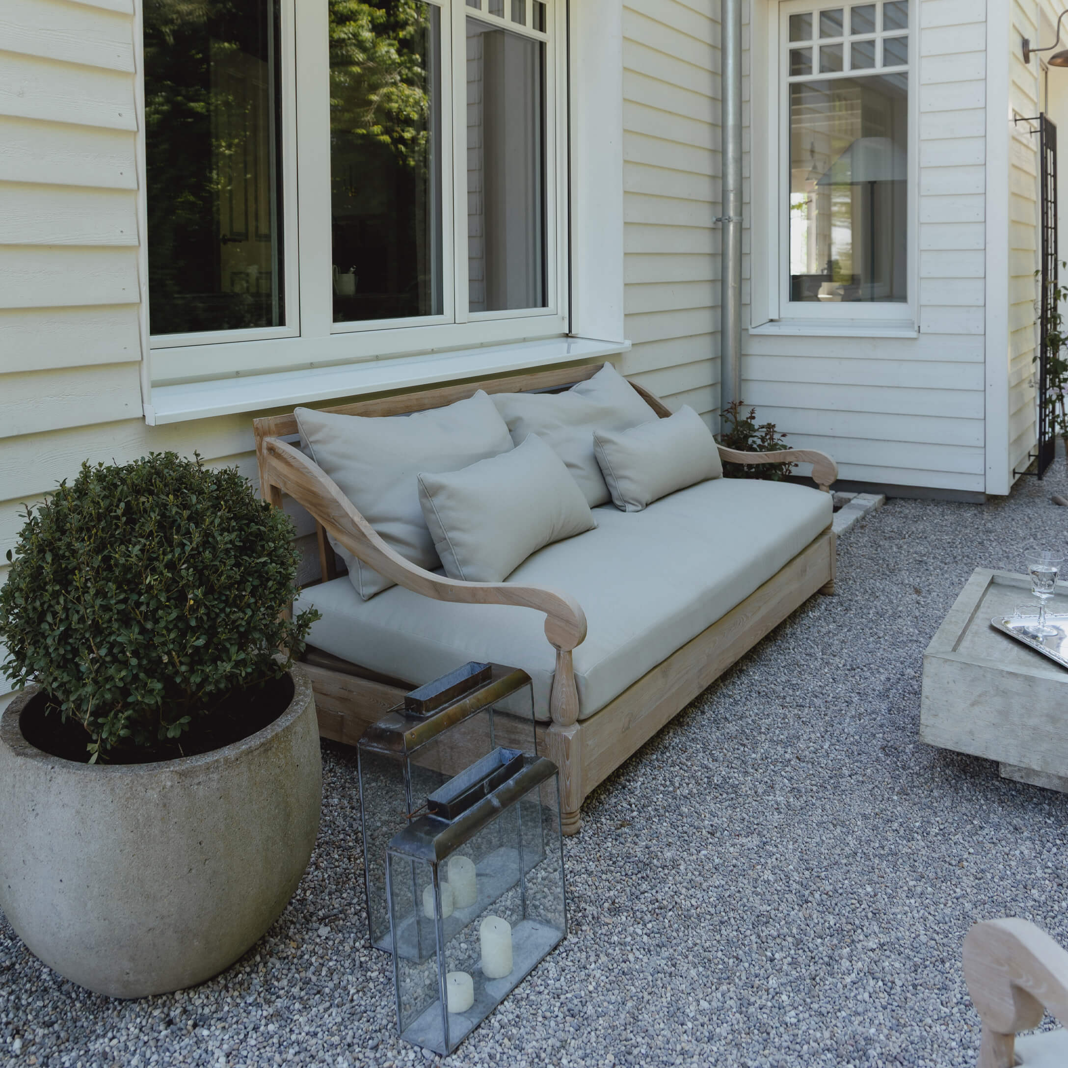 Drewniana sofa ogrodowa z jasnoszarymi poduszkami na żwirowym patio przy białym domu