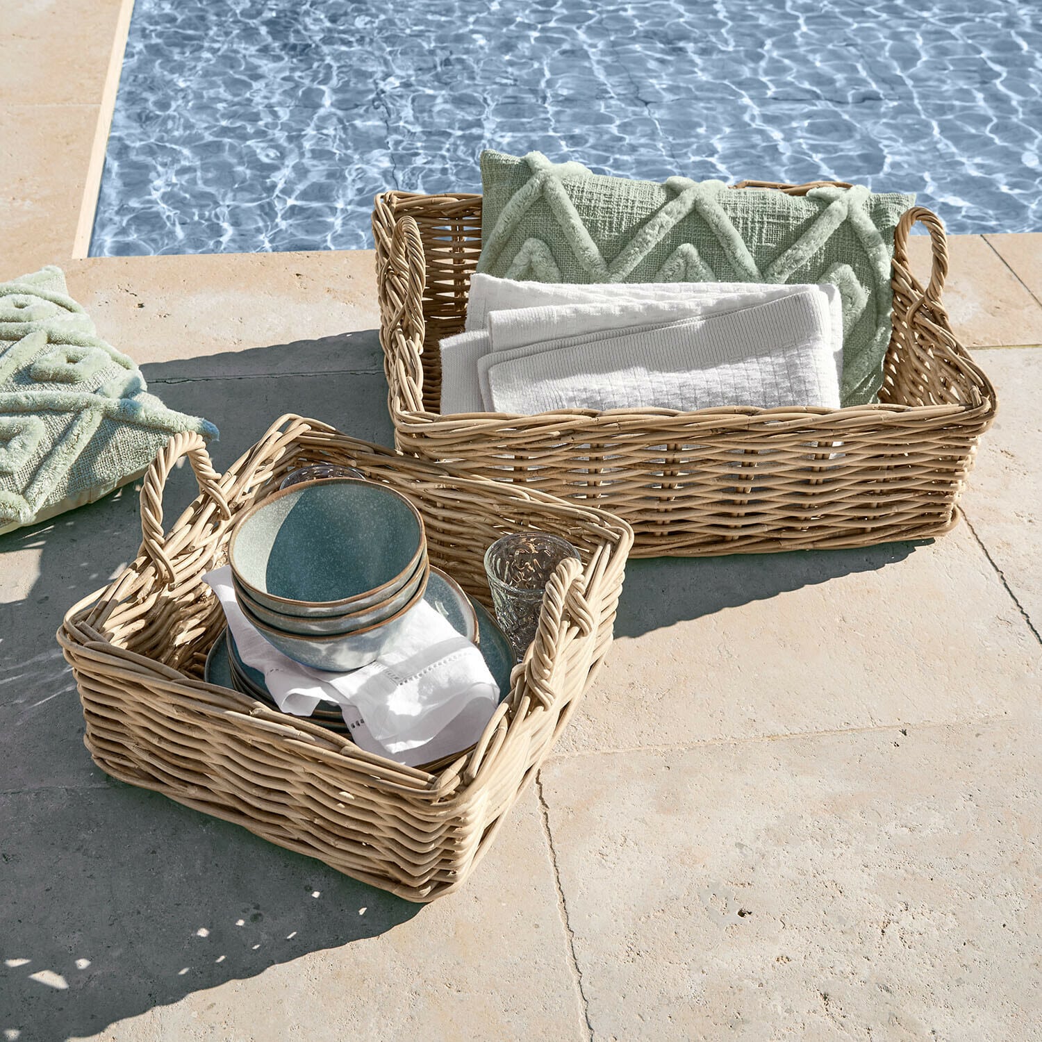 Dwa wiklinowe kosze z uchwytami, wypełnione zielonymi poduszkami i białymi ręcznikami przy basenie