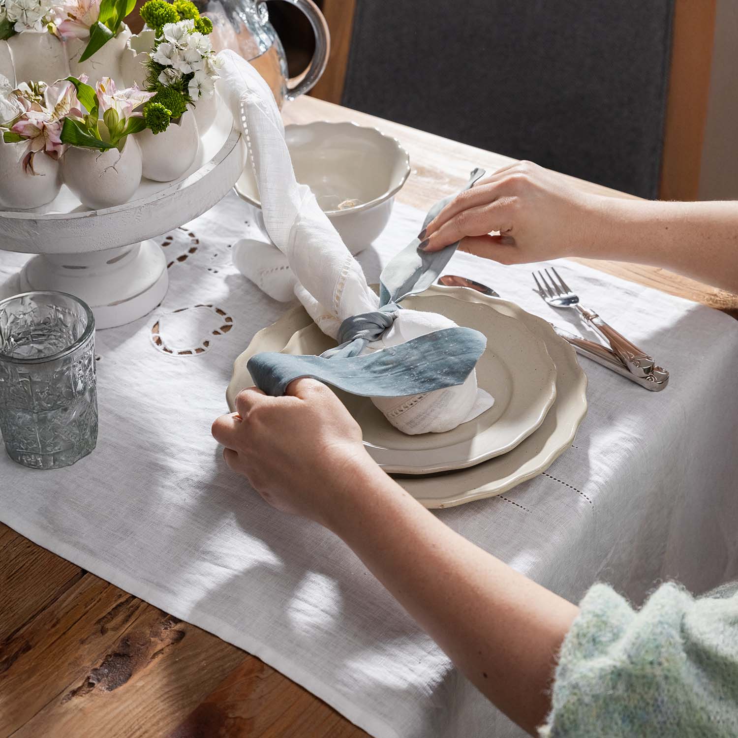 Ręce układające lniany biały obrus i serwetkę z niebieską wstążką na drewnianym stole z porcelanowymi talerzami