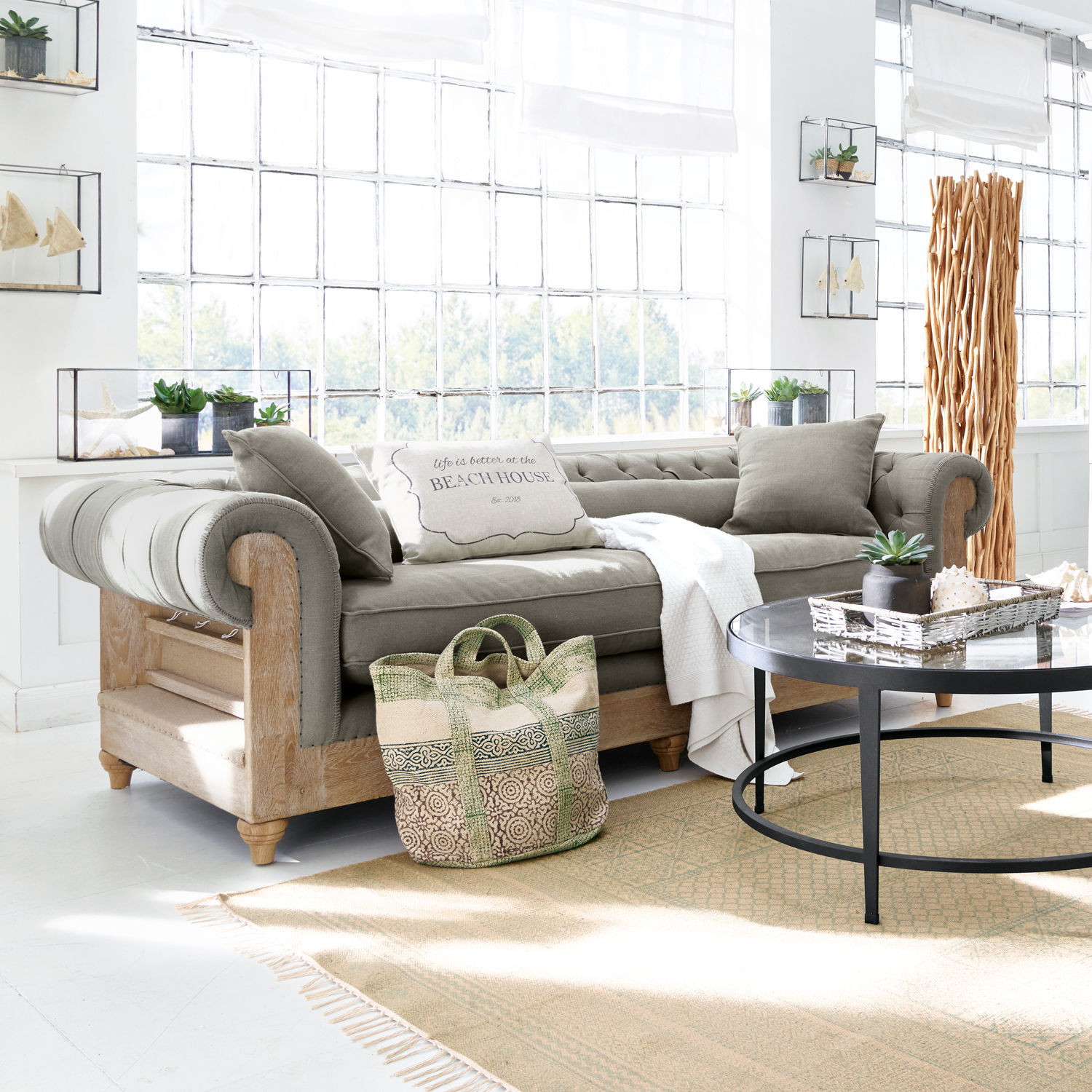 Sofa Kanani Szara sofa z drewnianą ramą w jasnym salonie z dekoracjami i oknami, styl skandynawski