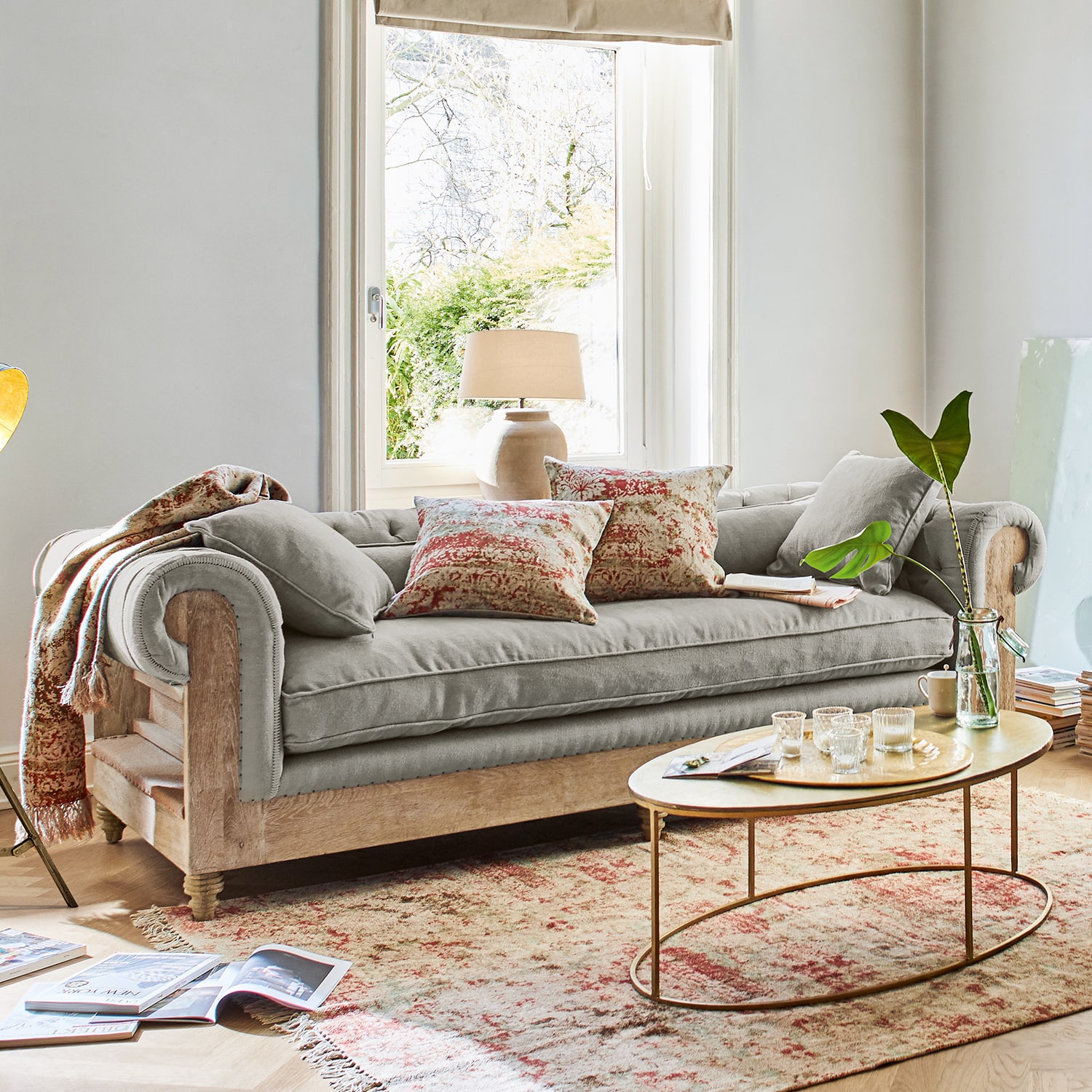 Szara sofa z drewnianą ramą i dekoracyjnymi poduszkami w jasnym salonie z oknem