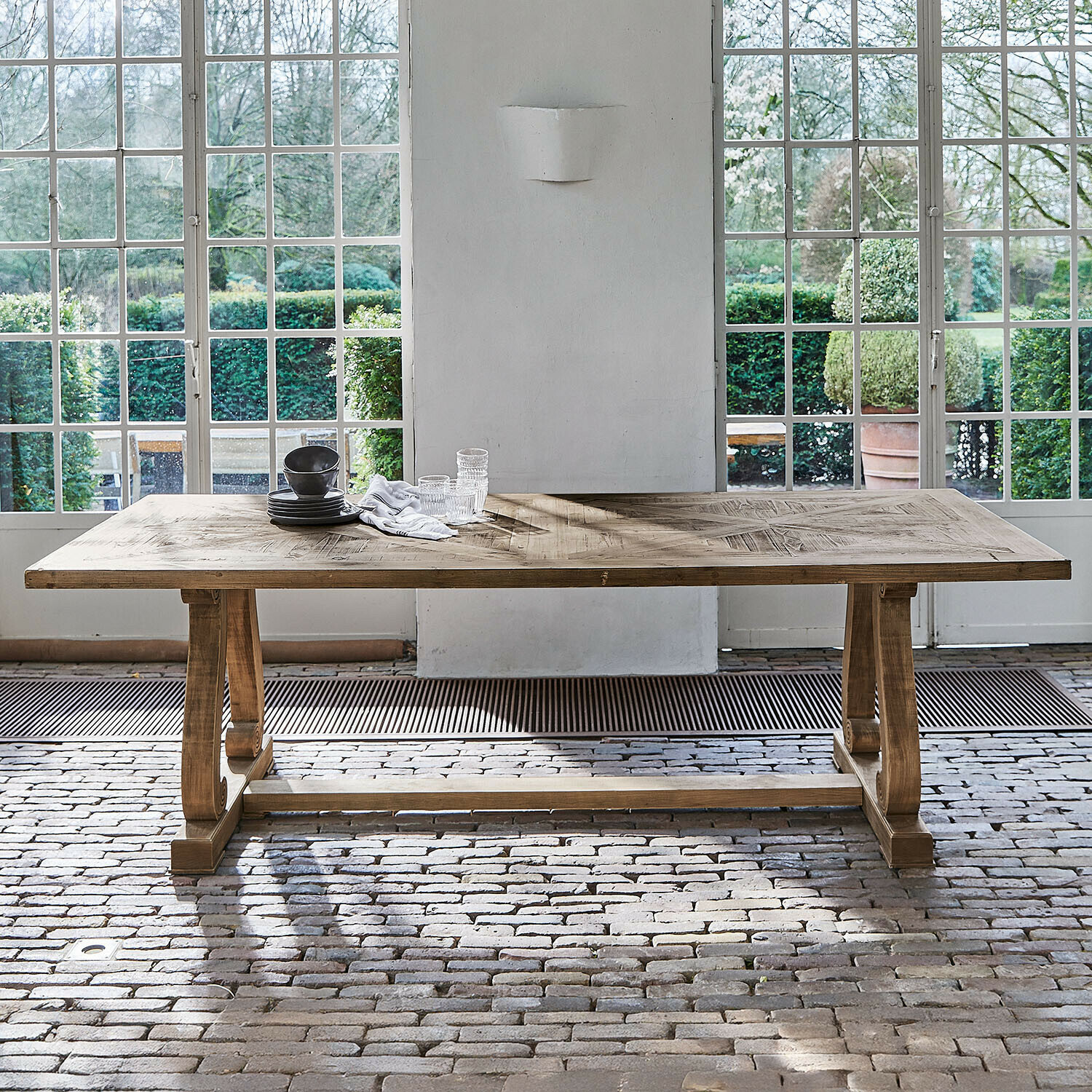 Duży drewniany stół jadalniany w naturalnym kolorze z ozdobnym blatem na tle dużych okien i ceglanej podłogi