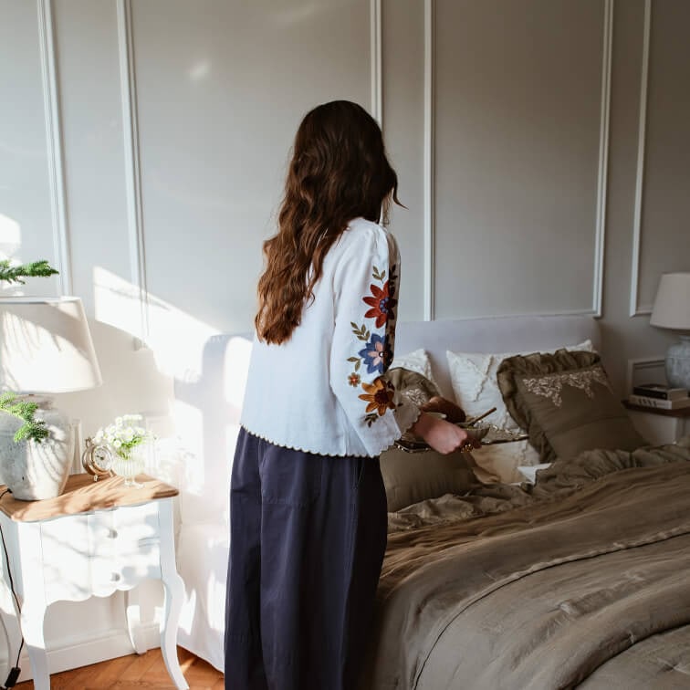 Kobieta w białej bluzce z kolorowym haftem na rękawach w sypialni z brązową pościelą i lampką na stoliku