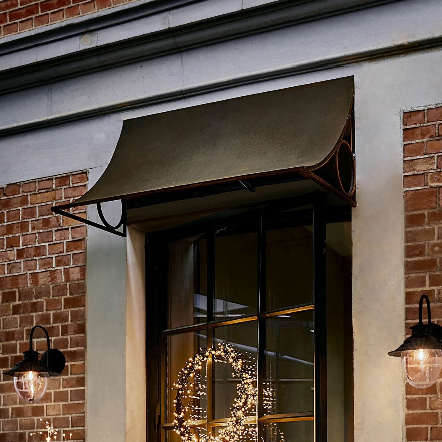 Metalowy daszek w kolorze oliwkowym nad oknem z czarną ramą i lampami ściennymi
