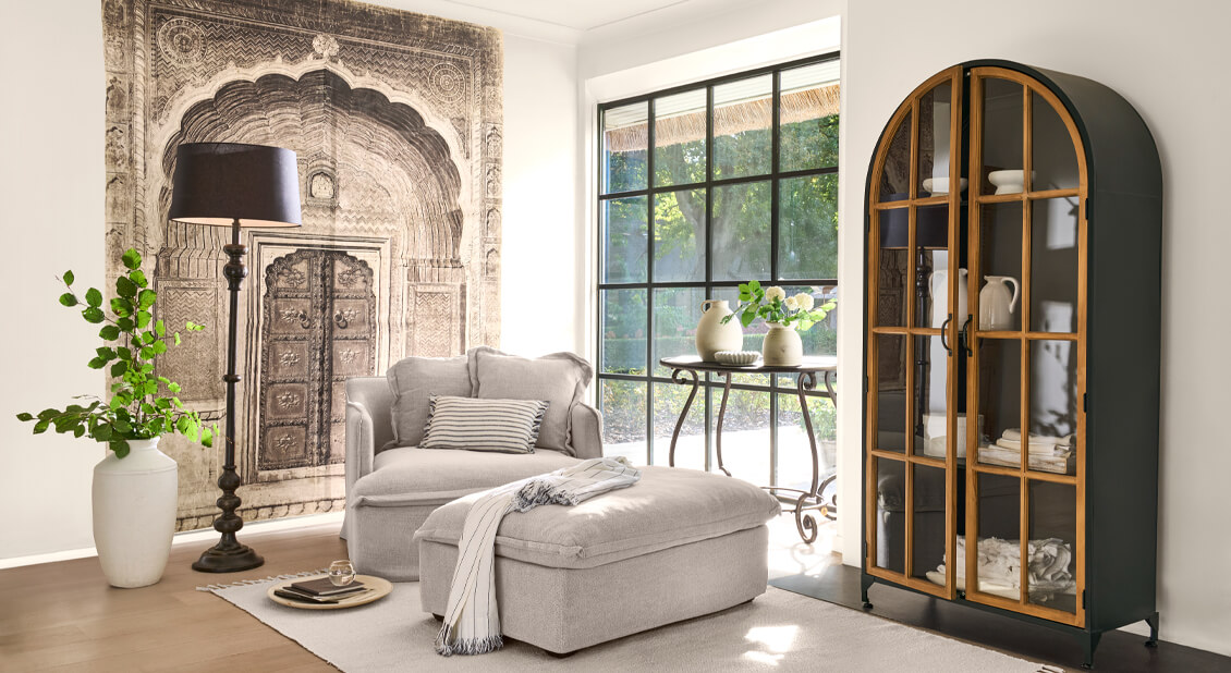 Przytulny salon z beżowym fotelem i pufą, metalową szafką z drewnianymi drzwiami i dekoracją ścienną w stylu orientalnym