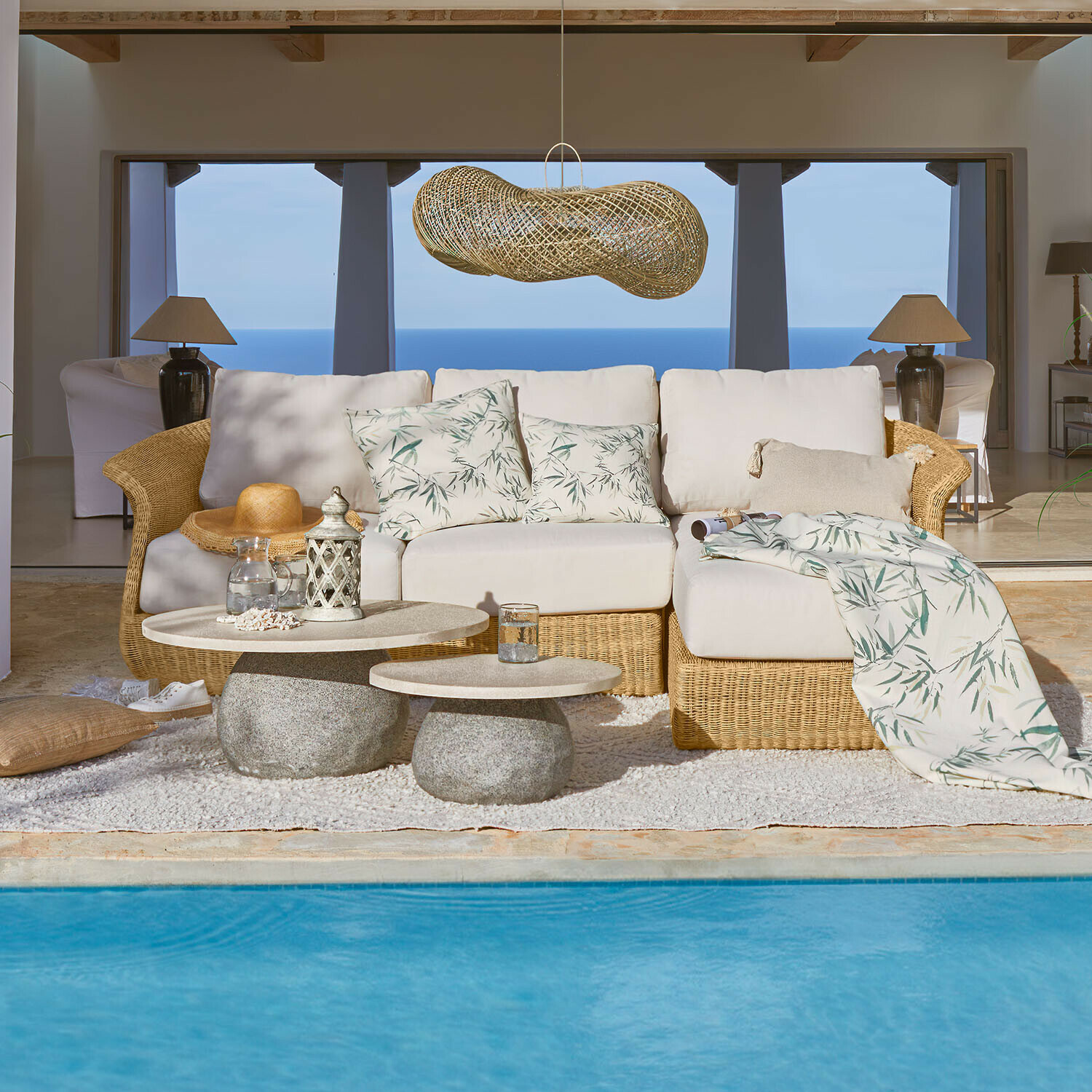 Komfortowa sofa z rattanu i jasnymi poduszkami z motywem liści przy basenie z widokiem na morze