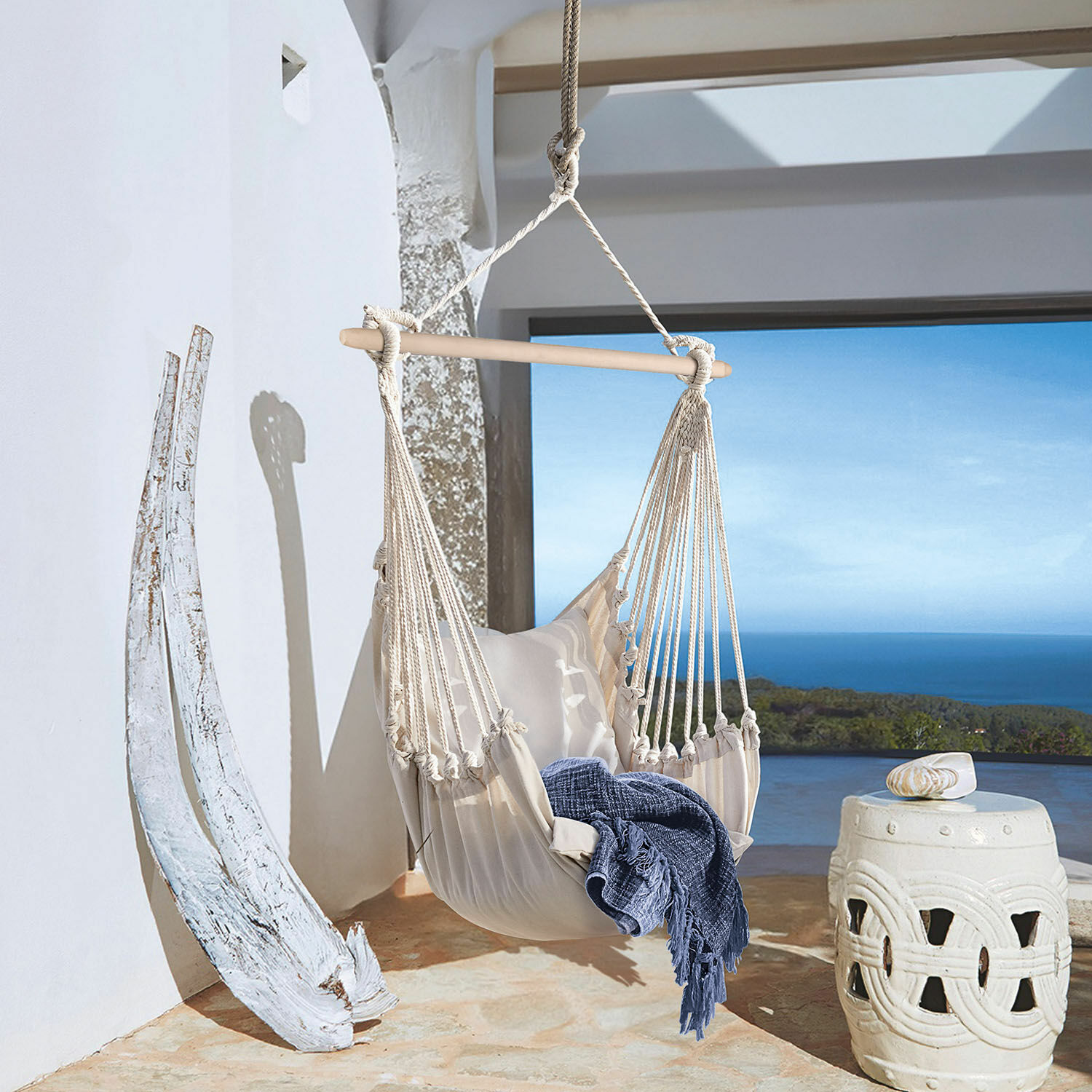 Fotel wiszący Leano Beżowy hamak z bawełny z drewnianą poprzeczką i niebieskim kocem na tarasie z widokiem na morze
