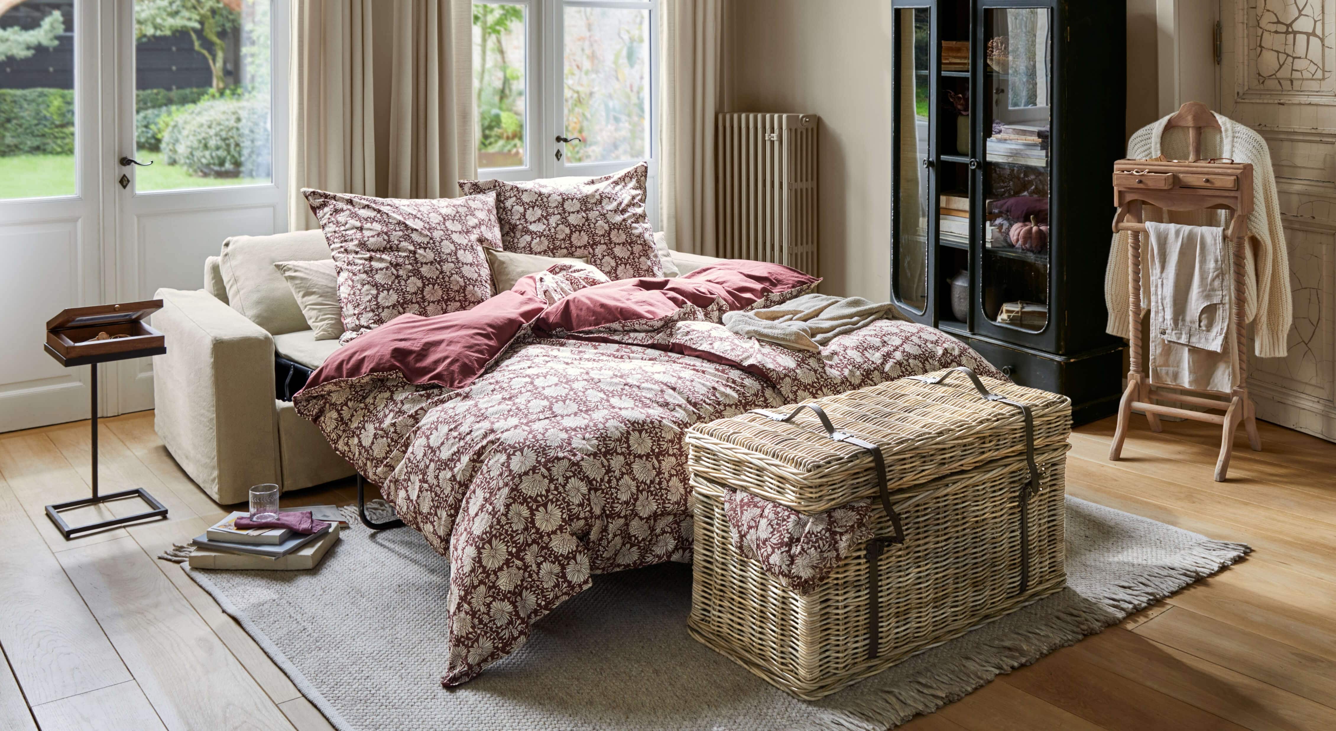 Rozkładana sofa z beżową tapicerką i bordową pościelą w kwiatowy wzór w jasnym salonie
