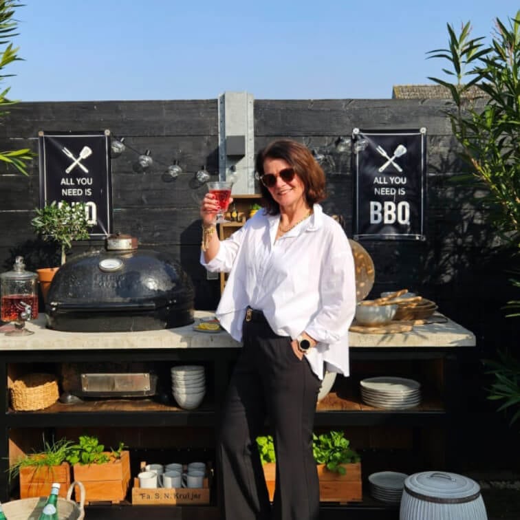 Kobieta w białej koszuli i czarnych spodniach stoi przy zewnętrznym grillu z napisem BBQ na tle czarnej ściany