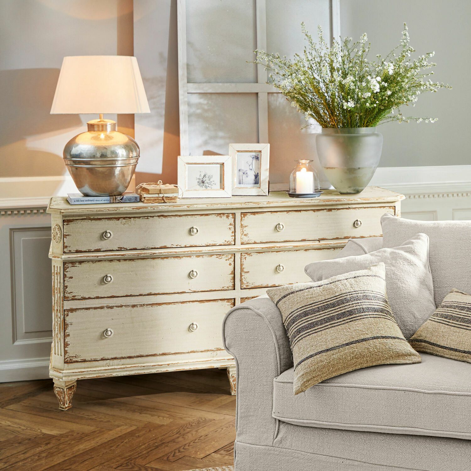 Komoda Commeren Kremowa drewniana komoda w stylu vintage z lampą, ramkami i dekoracjami w salonie z beżową sofą