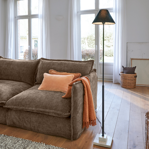 Brązowa sofa z miękką tkaniną, pomarańczowa poduszka i pled, lampa podłogowa z czarnym abażurem i marmurową podstawą