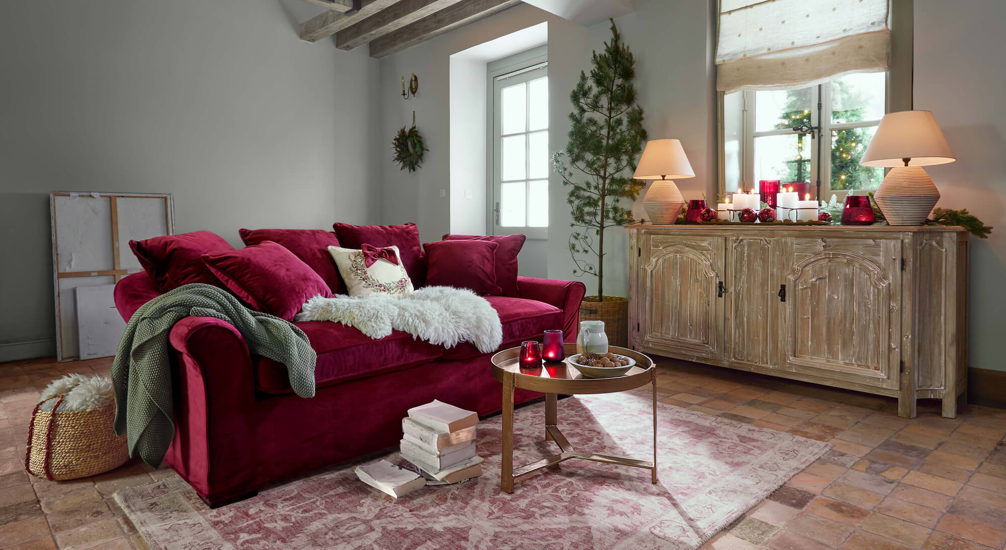 Czerwony welurowy sofa z poduszkami i zielonym kocem w przytulnym salonie z drewnianą komodą i dekoracjami świątecznymi