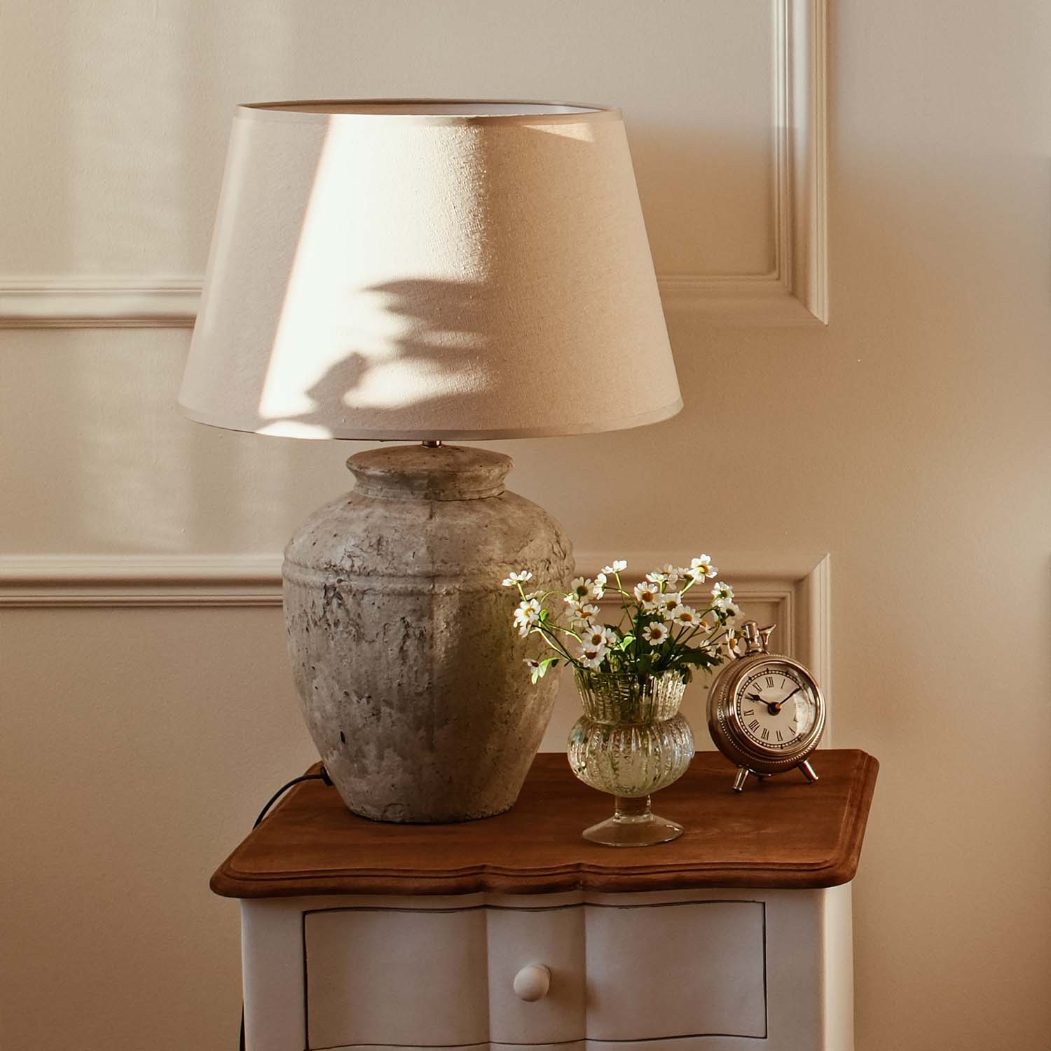 Lampka stołowa z beżowym abażurem i ceramiczną podstawą w stylu vintage na drewnianym stoliku z kwiatami i zegarem