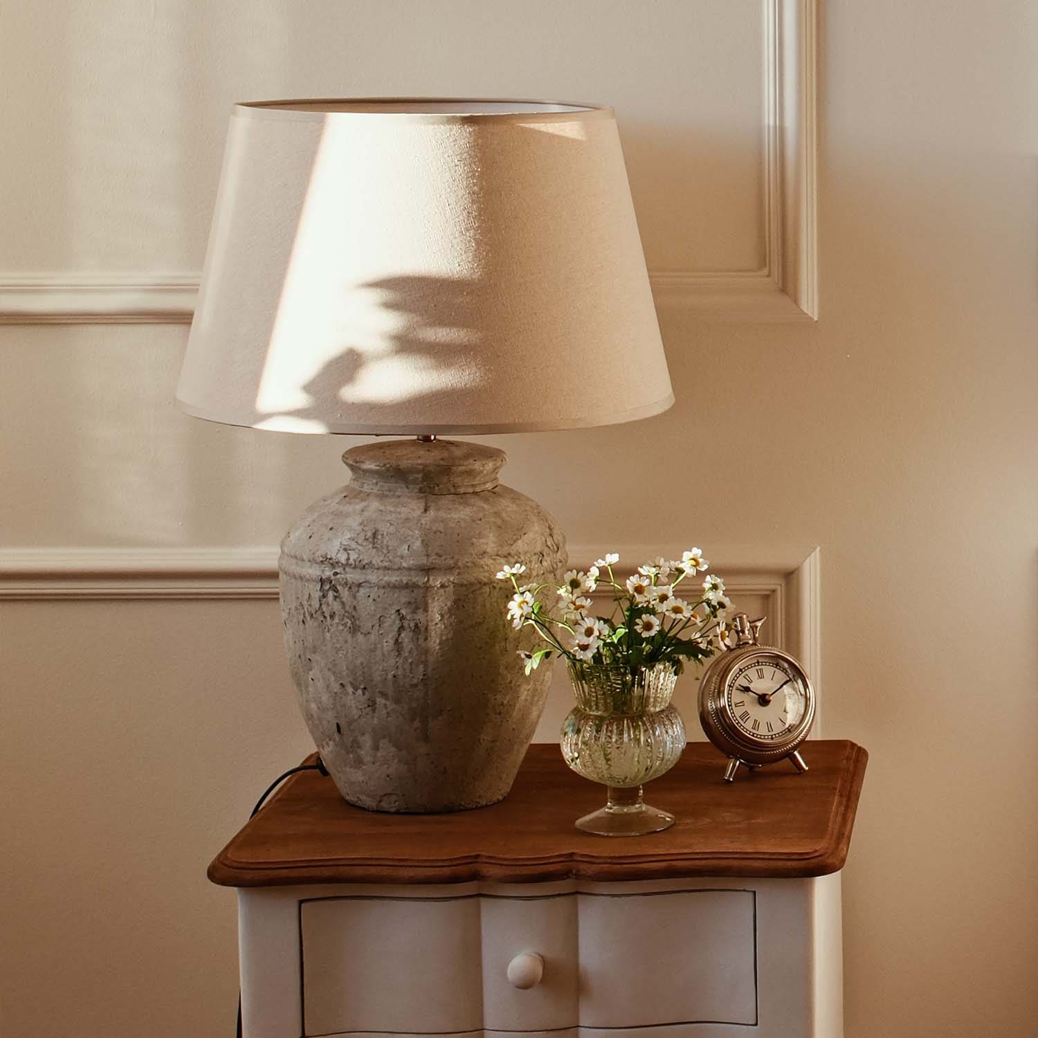 Lampka stołowa z beżowym abażurem i ceramiczną podstawą w stylu vintage na drewnianym stoliku z kwiatami i zegarem