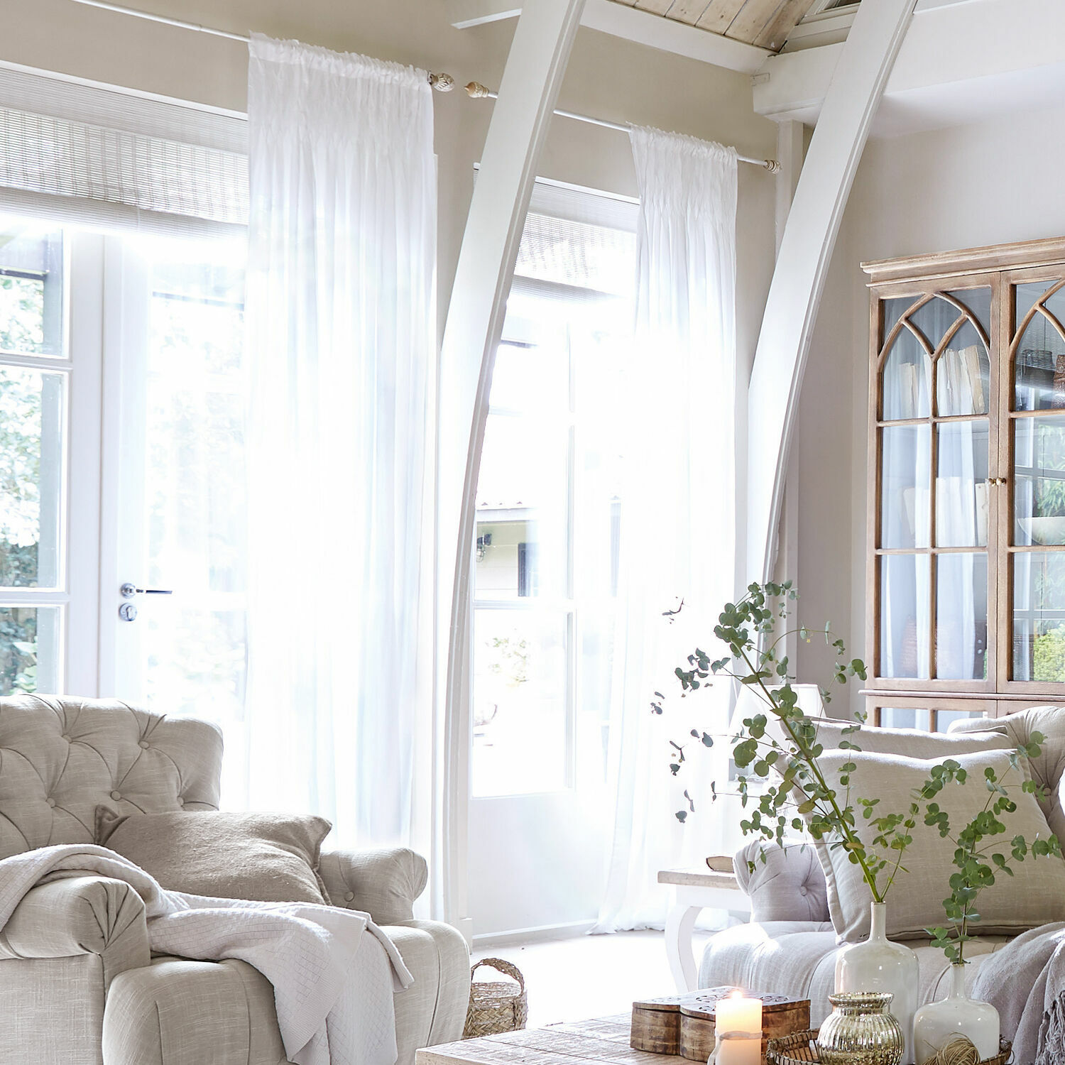 Firana Otilie Jasny salon z beżową tapicerką, białymi zasłonami i drewnianą witryną w stylu vintage