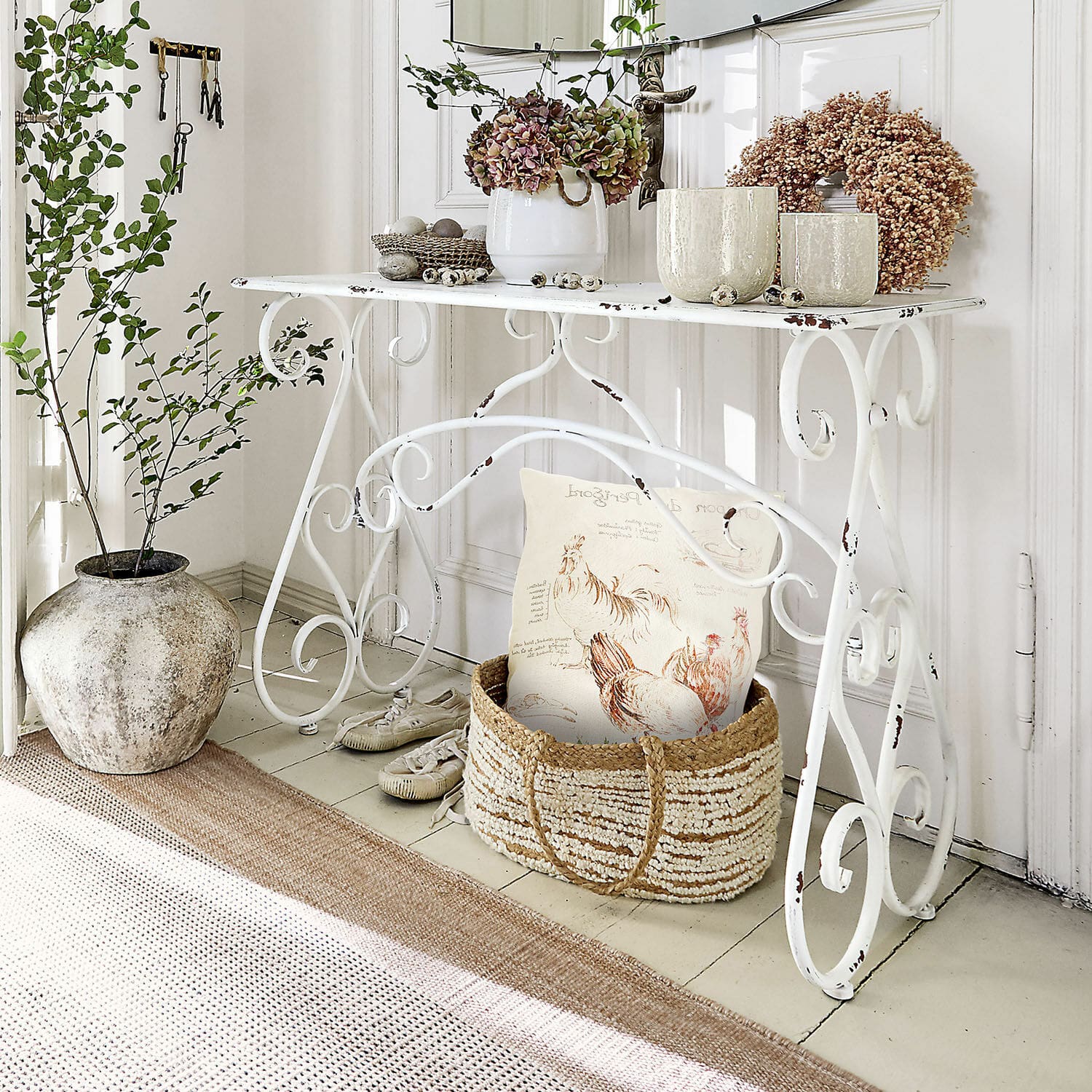 Biały metalowy stolik z dekoracyjnymi roślinami i wiklinowym koszem z poduszką w stylu rustykalnym