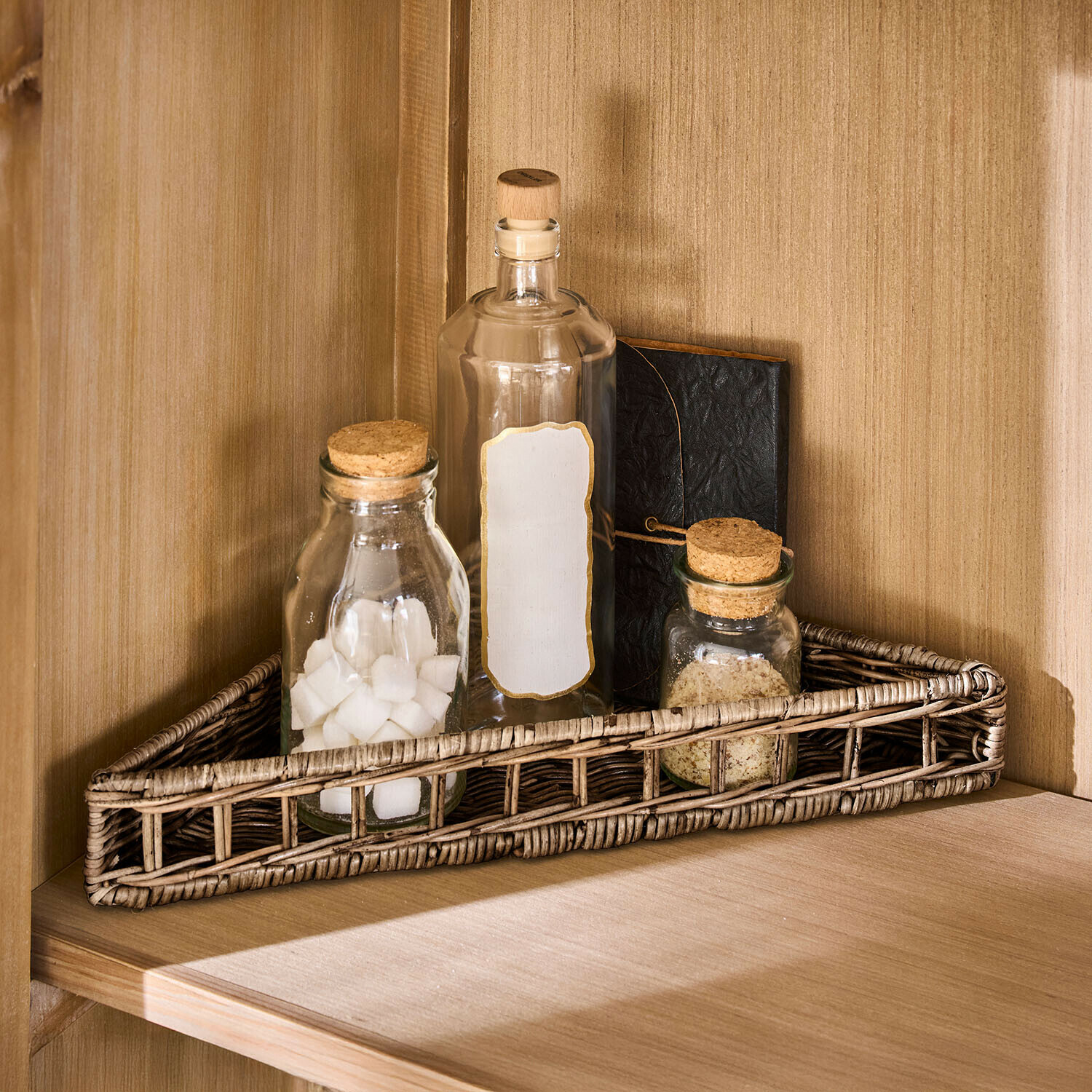 Trójkątny koszyk z rattanu w naturalnym kolorze z trzema szklanymi butelkami i notesem na drewnianej półce