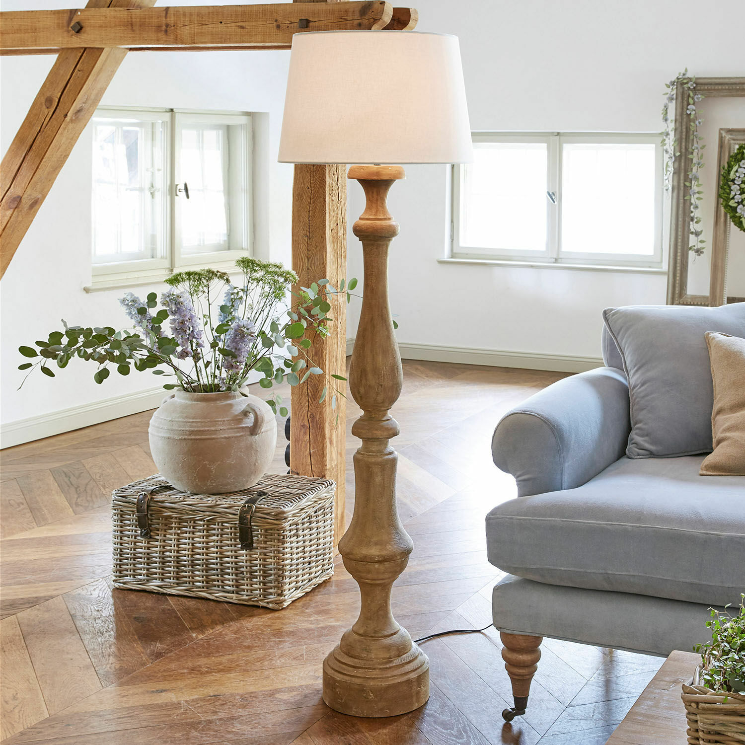 Lampa stojąca Etienne Drewniana lampa podłogowa z białym abażurem w jasnym salonie z niebieską sofą i wiklinowym koszem