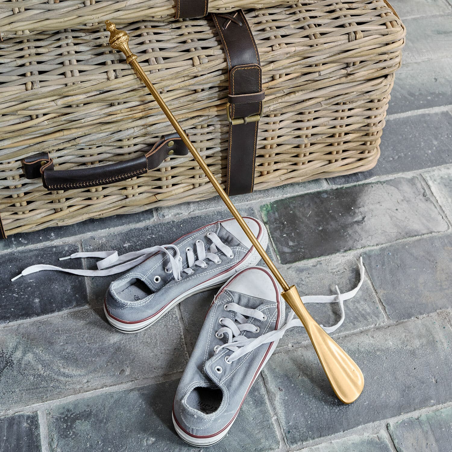 Łyżka do butów Orveline Złota metalowa łyżka do butów na tle wiklinowego kosza i szarych tenisówek, akcesorium do obuwia