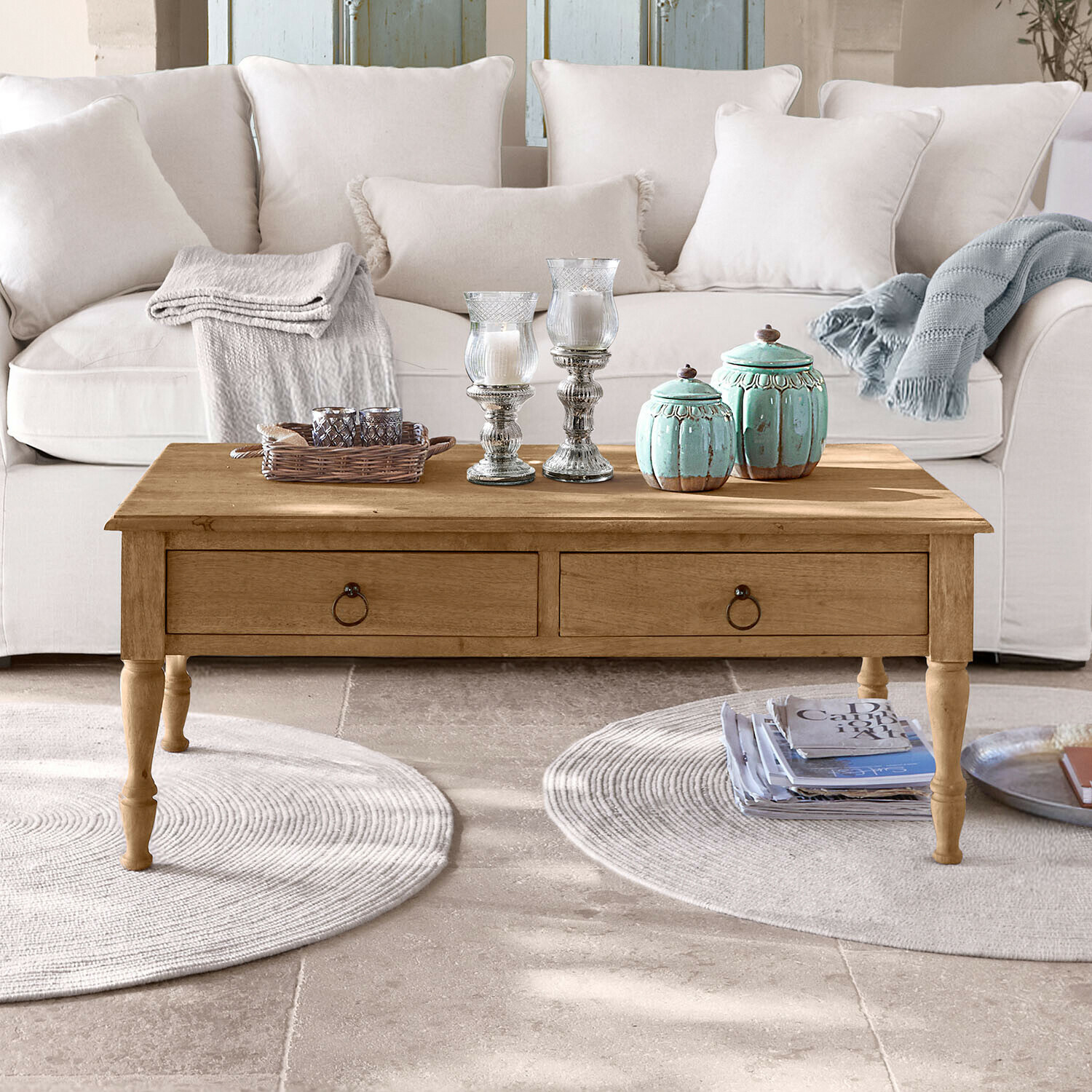 Drewniany stolik kawowy z szufladami na tle jasnej kanapy, dekoracje i pledy na blacie