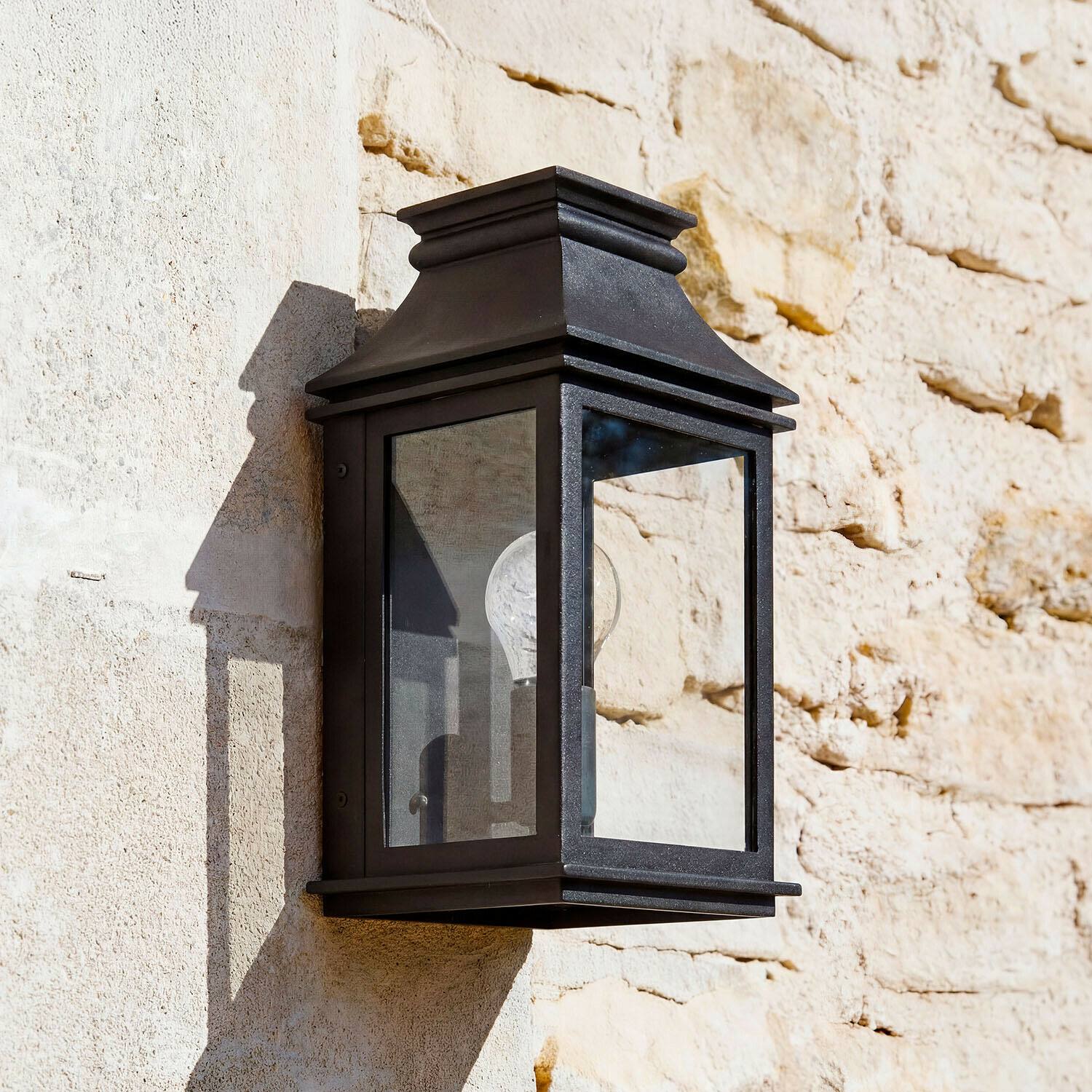 Kinkiet zewnętrzny Zélivarne Czarna metalowa lampa ścienna z przezroczystym szkłem na jasnej kamiennej ścianie, oświetlenie zewnętrzne