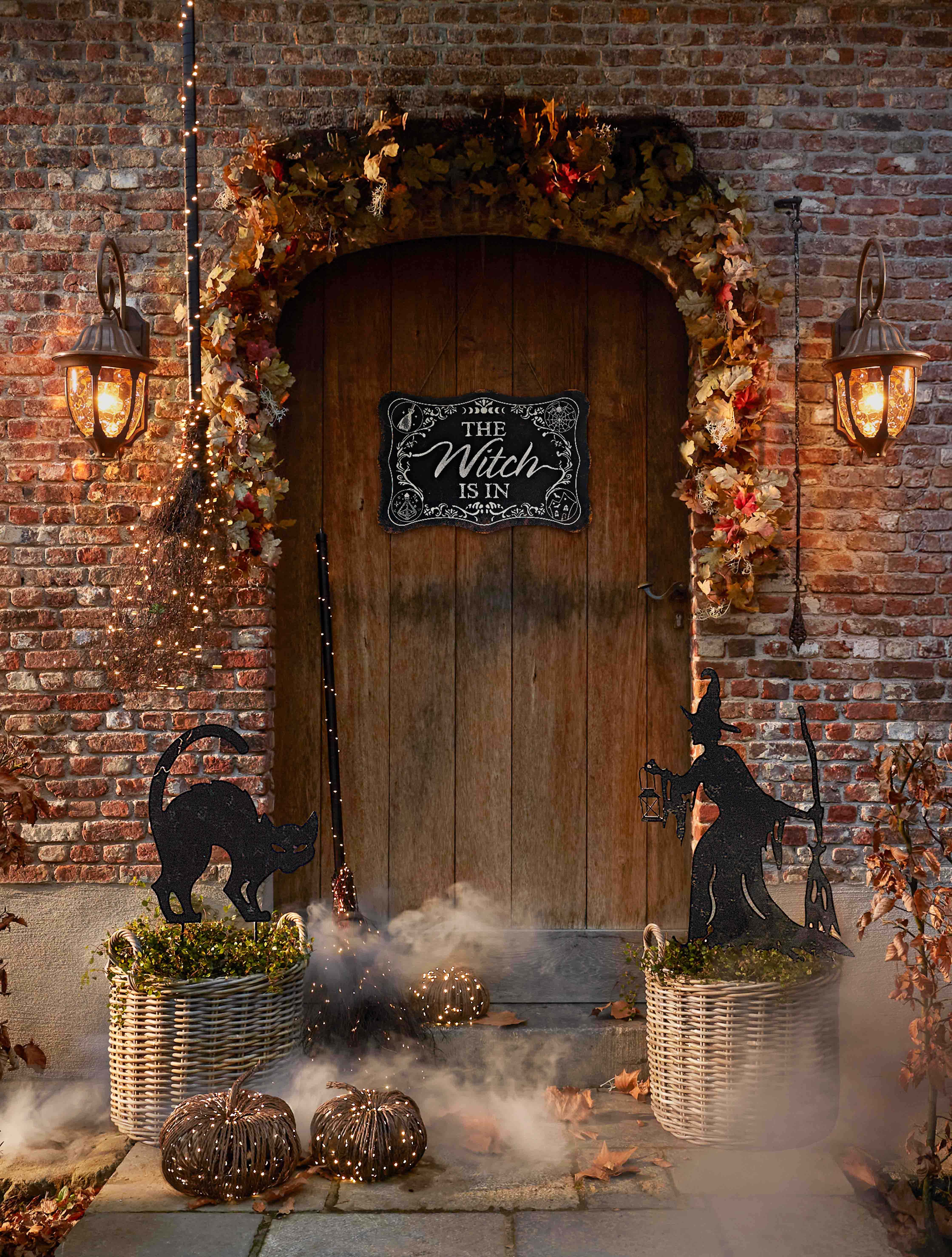 Dekoracja halloweenowa z czarownicą, czarnym kotem i lampionami przy drewnianych drzwiach z cegły