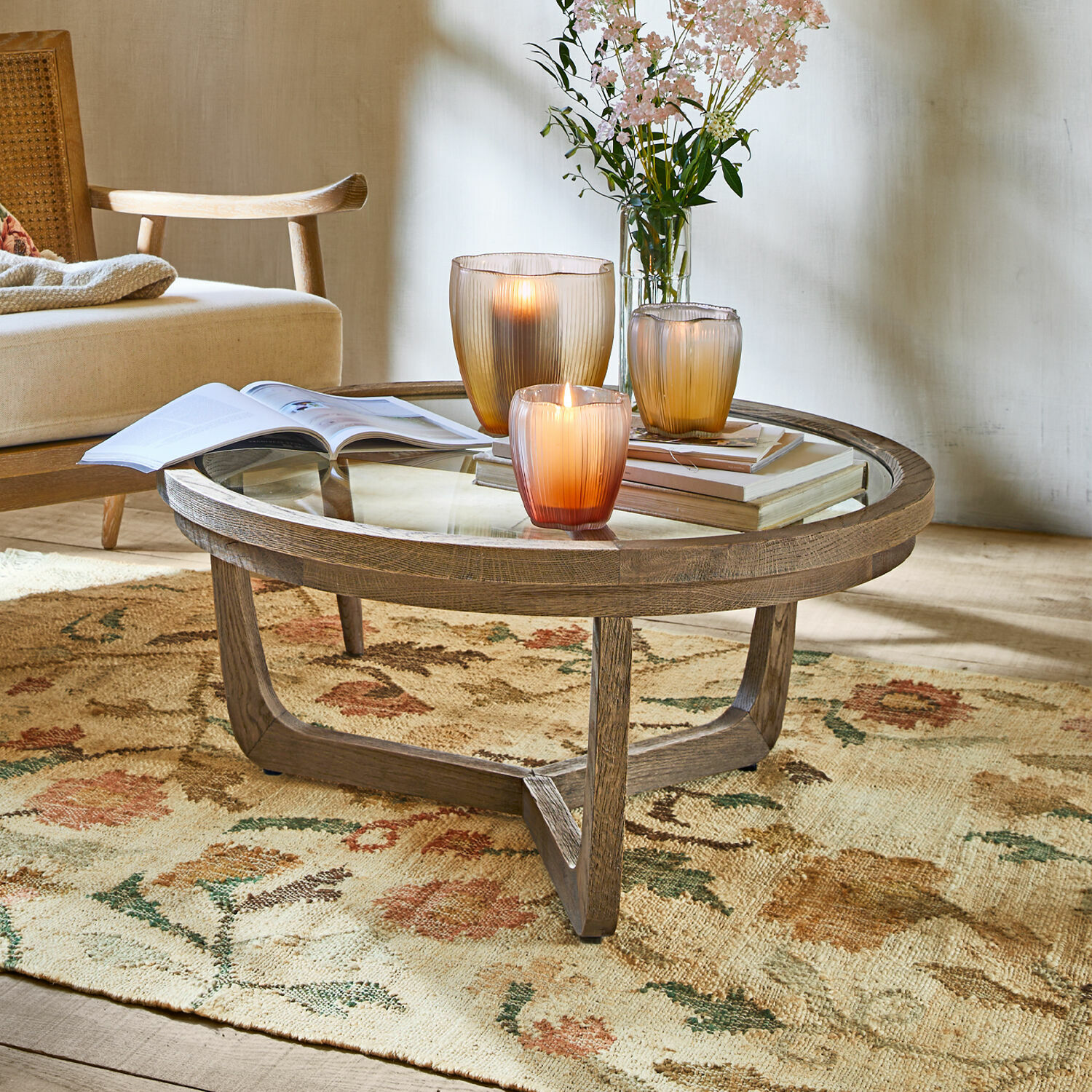 Okrągły stolik kawowy z drewna i szkła na kwiecistym dywanie, dekorowany świecami i książkami