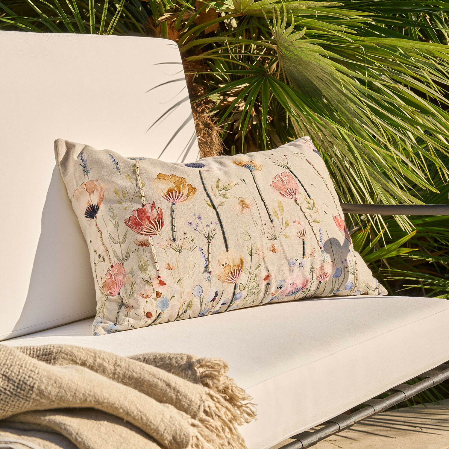 Beżowa poduszka dekoracyjna z kwiatowym wzorem na jasnym fotelu ogrodowym z kocem