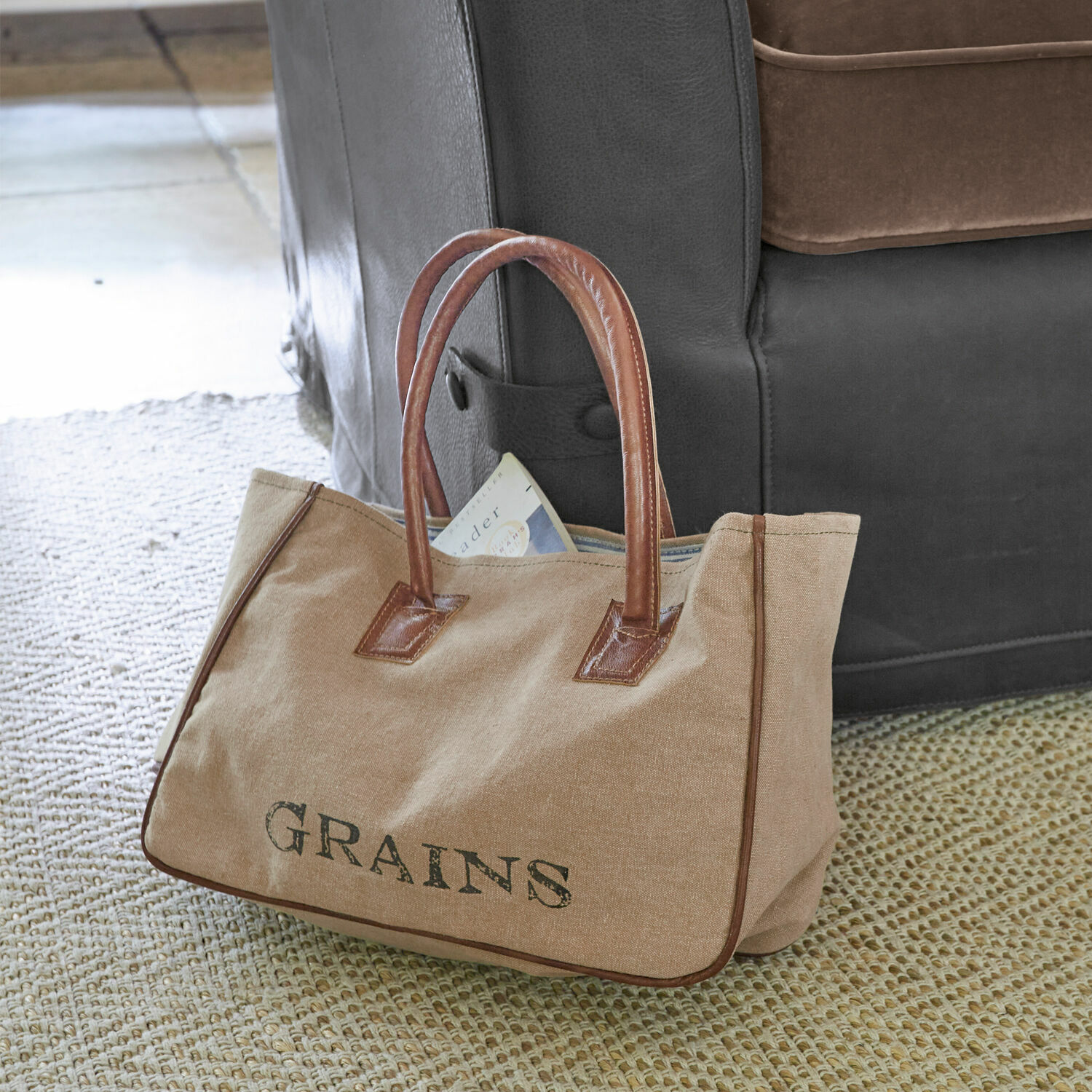 Torba Grains Beżowa torba z materiału z brązowymi skórzanymi uchwytami i napisem GRAINS na dywanie przy kanapie