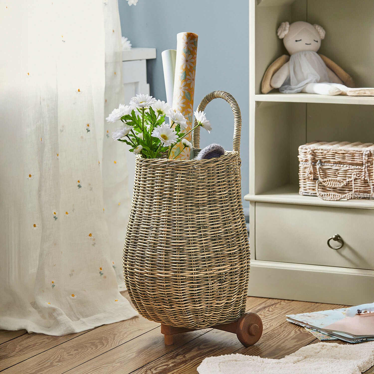Wiklinowy kosz na kółkach z uchwytem, wypełniony białymi kwiatami i papierem dekoracyjnym, naturalny kolor