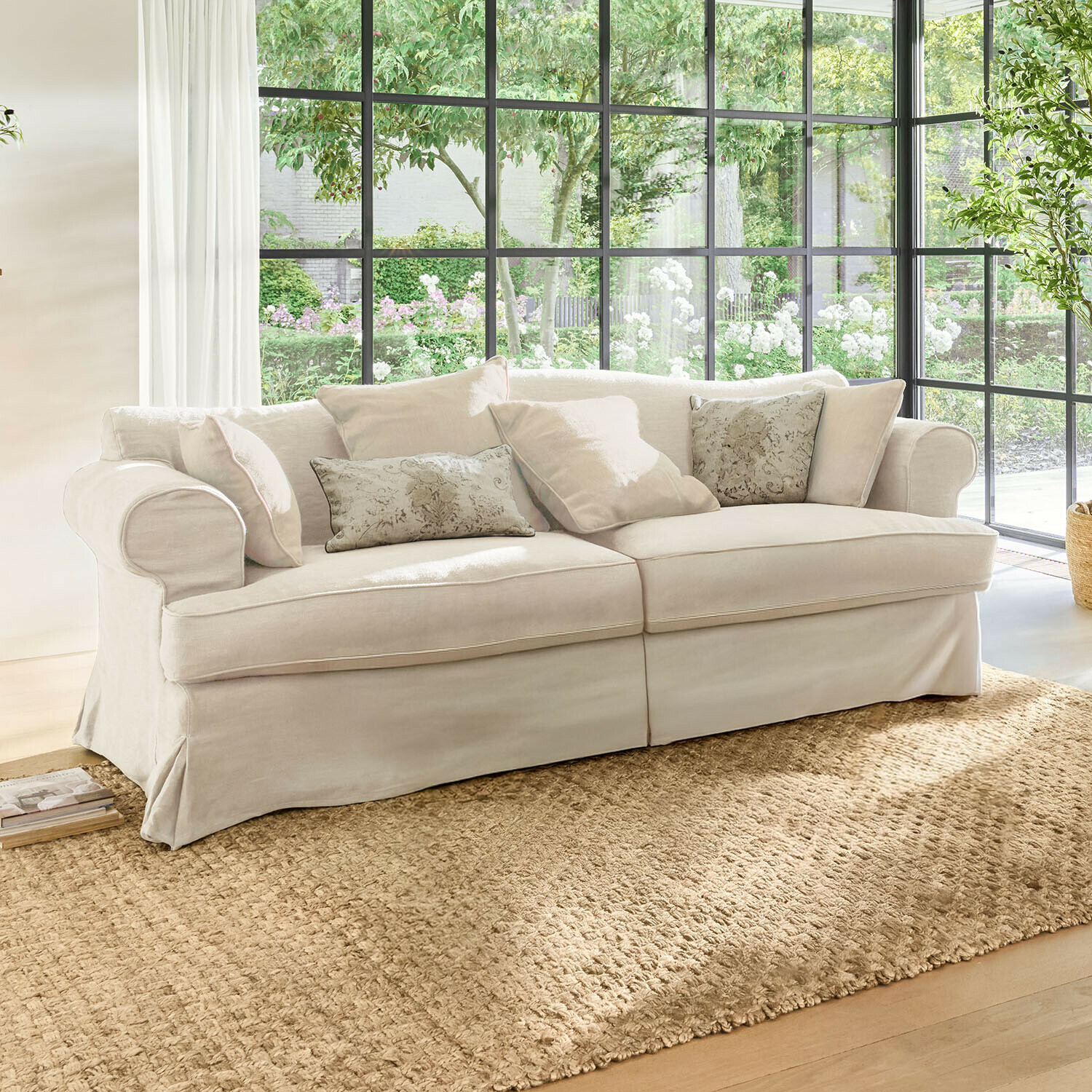 Sofa Dawnhurst Beżowa sofa z tkaniny z poduszkami dekoracyjnymi na naturalnym dywanie w jasnym salonie z dużymi oknami