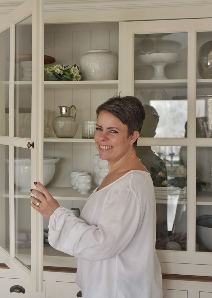 Kobieta otwierająca kremową szafkę kuchenną z porcelanowymi naczyniami i dekoracjami w tle