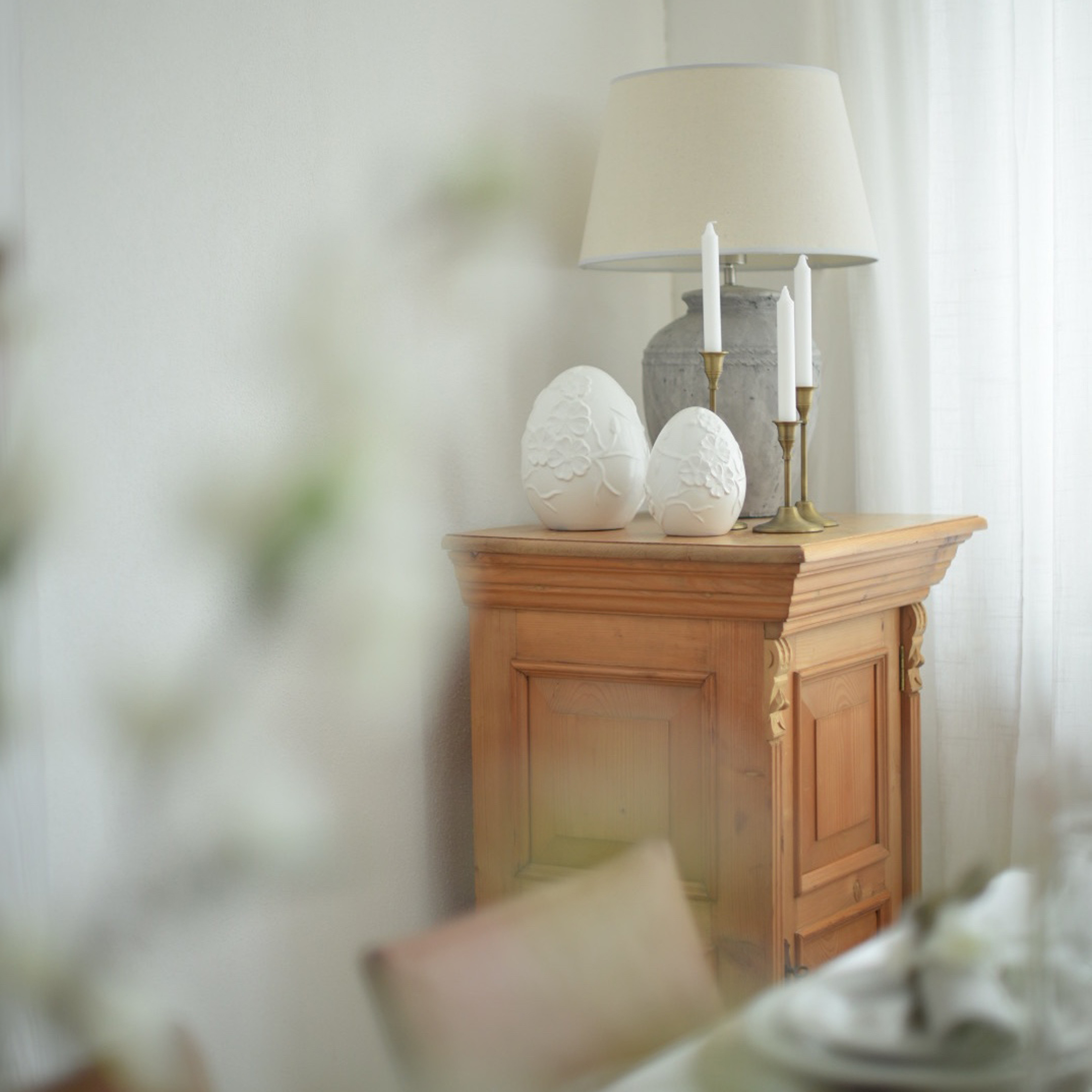 Drewniana szafka z lampą, świecami i białymi ceramicznymi dekoracjami w kształcie jajek w jasnym wnętrzu