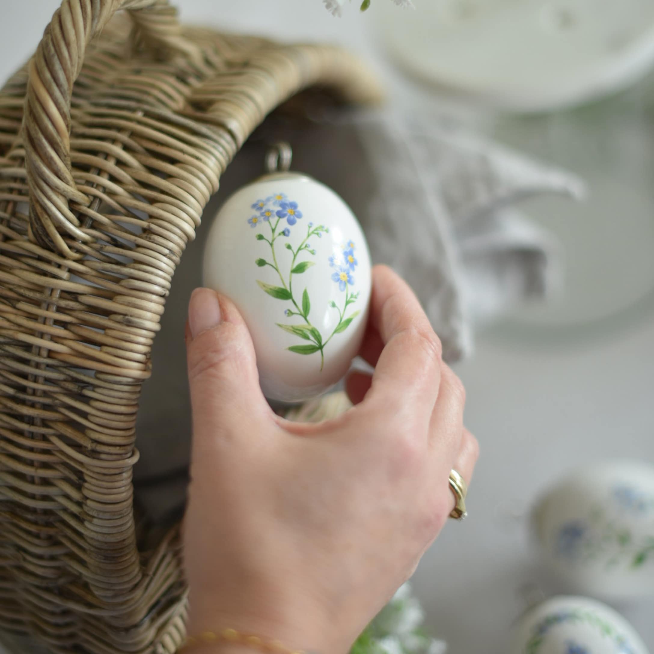 Ręka trzymająca ceramiczną białą ozdobę wielkanocną z niebieskimi kwiatami obok wiklinowego koszyka